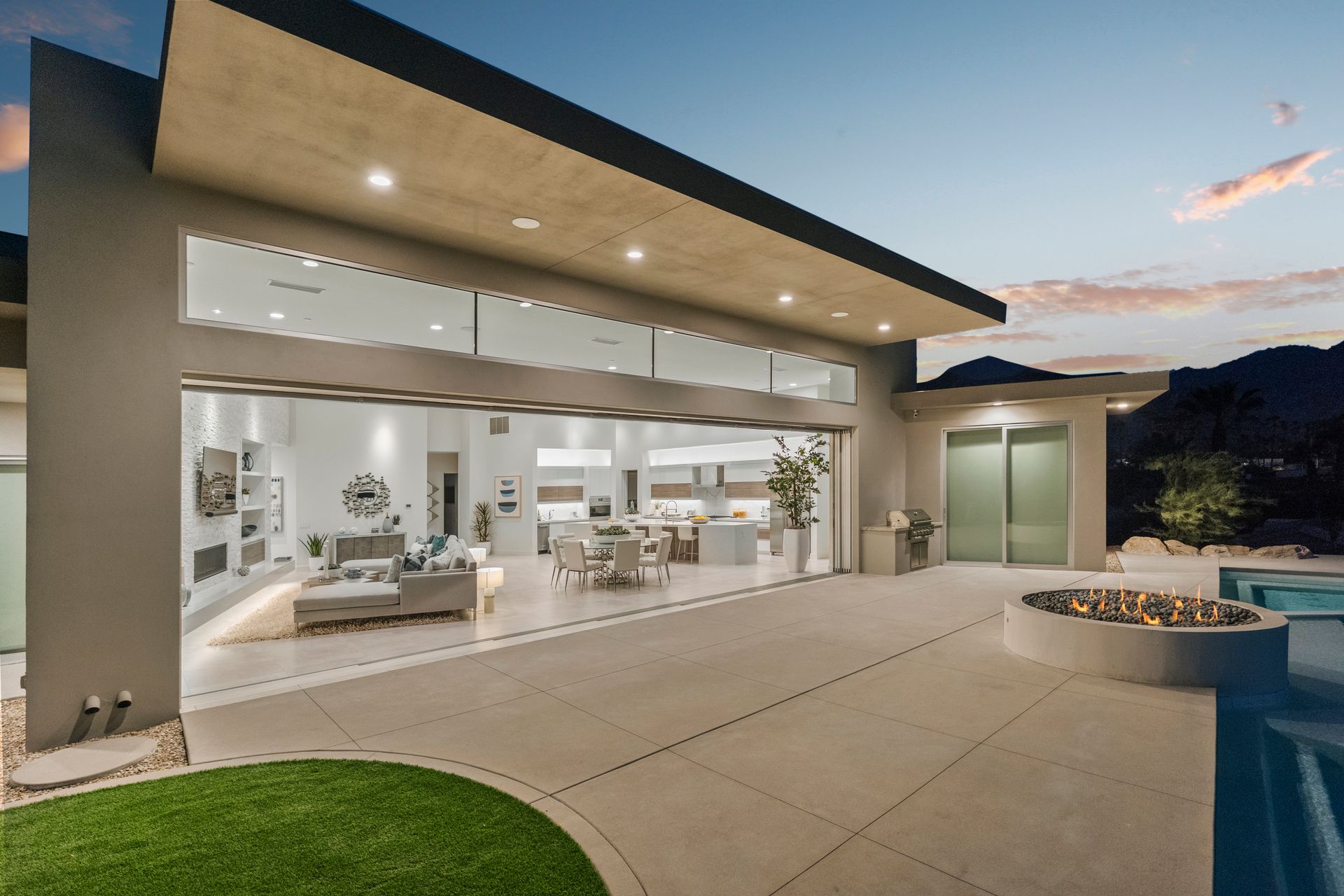 Modern patio with a fire pit and pool, opening into a spacious, illuminated living area at dusk.