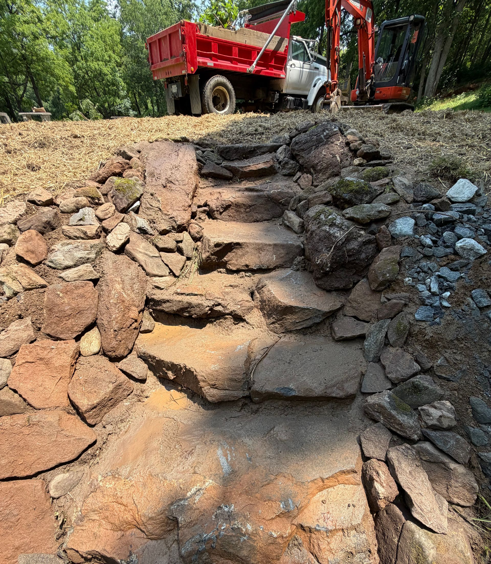Red dump truck and excavator beside a rough stone retaining wall in a wooded worksite.