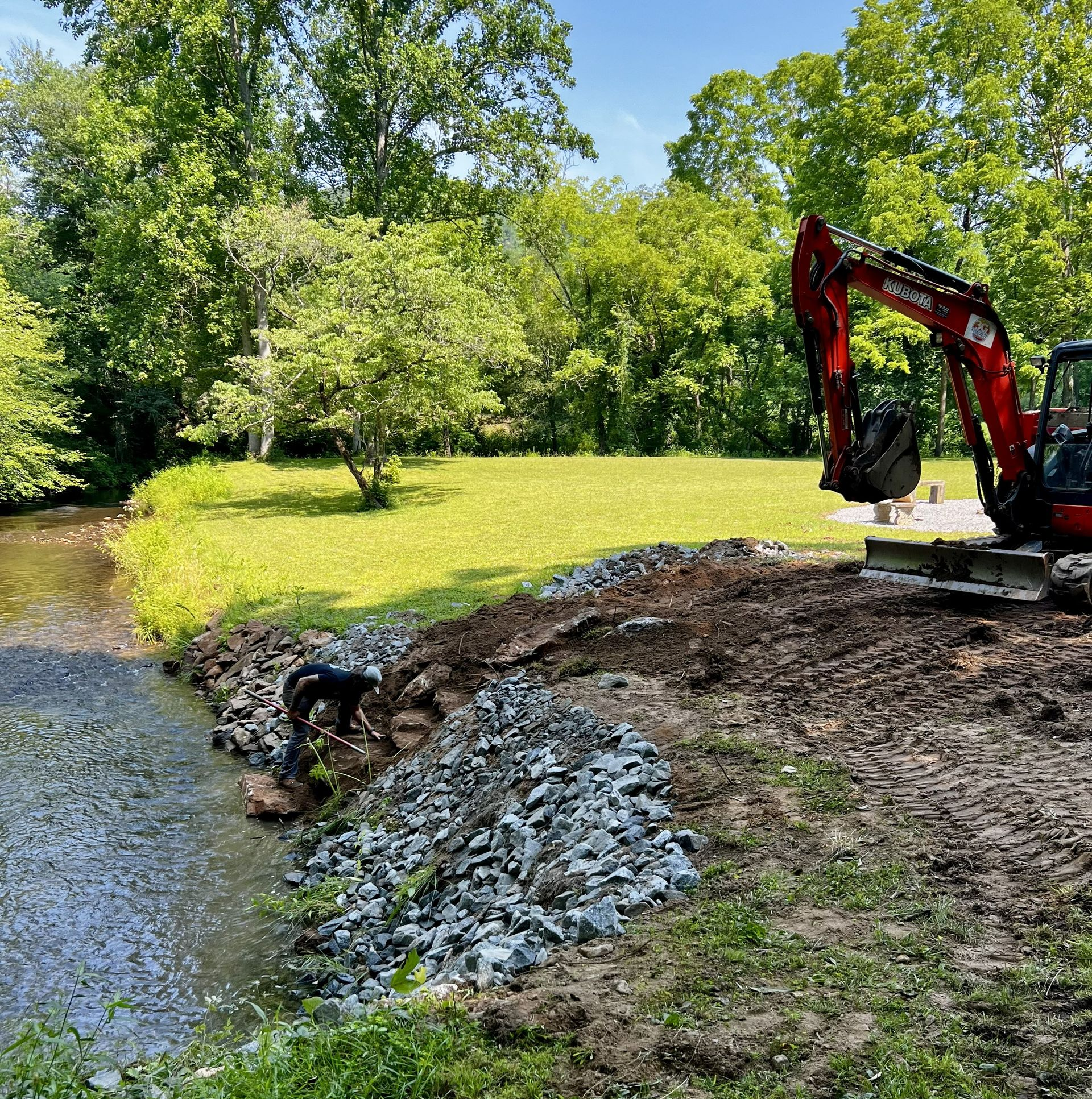 Red excavator beside a creek, moving rocks along a grassy riverbank in a wooded park