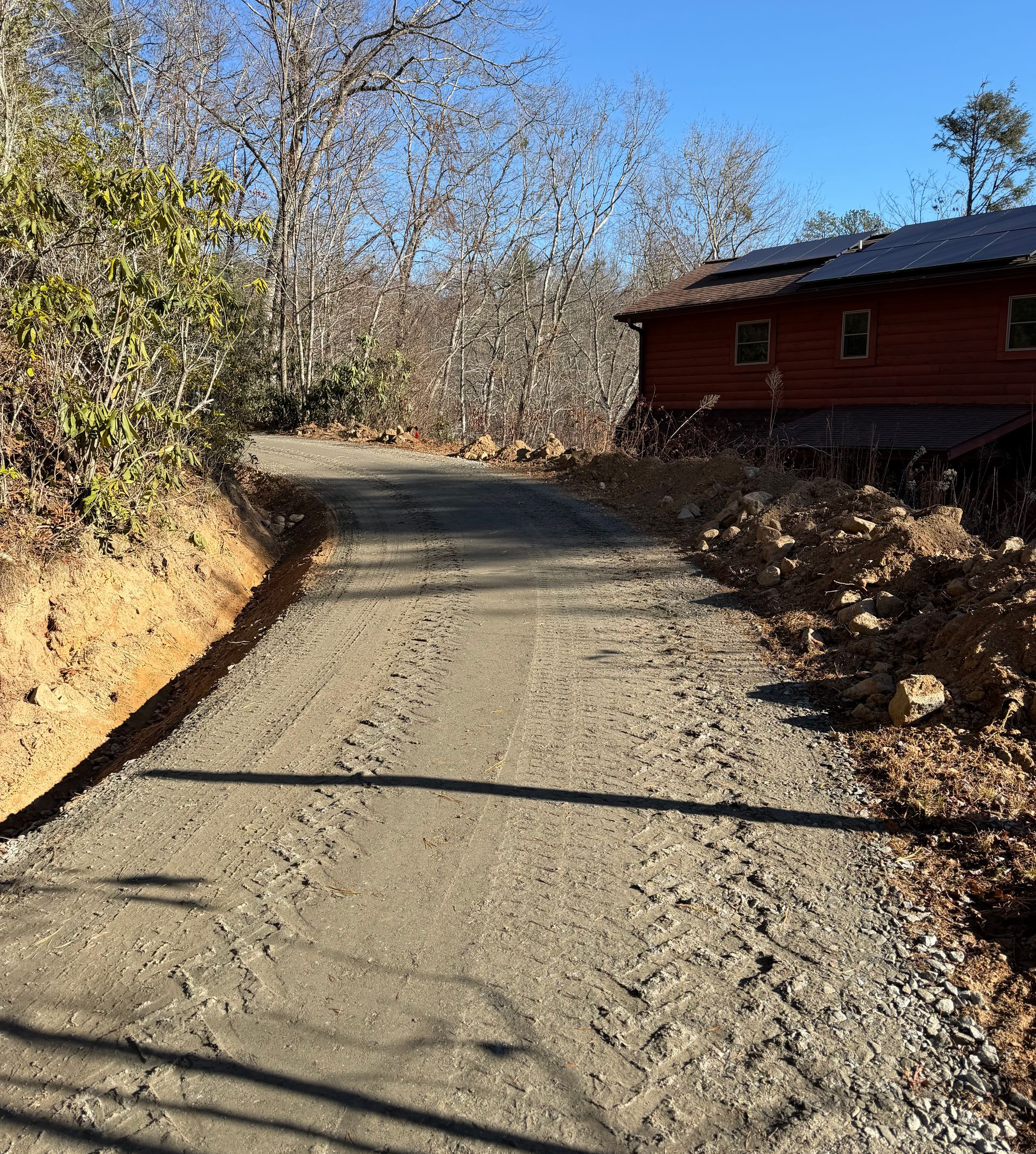 Dirt road curving uphill beside a red cabin in a wooded area under a clear blue sky