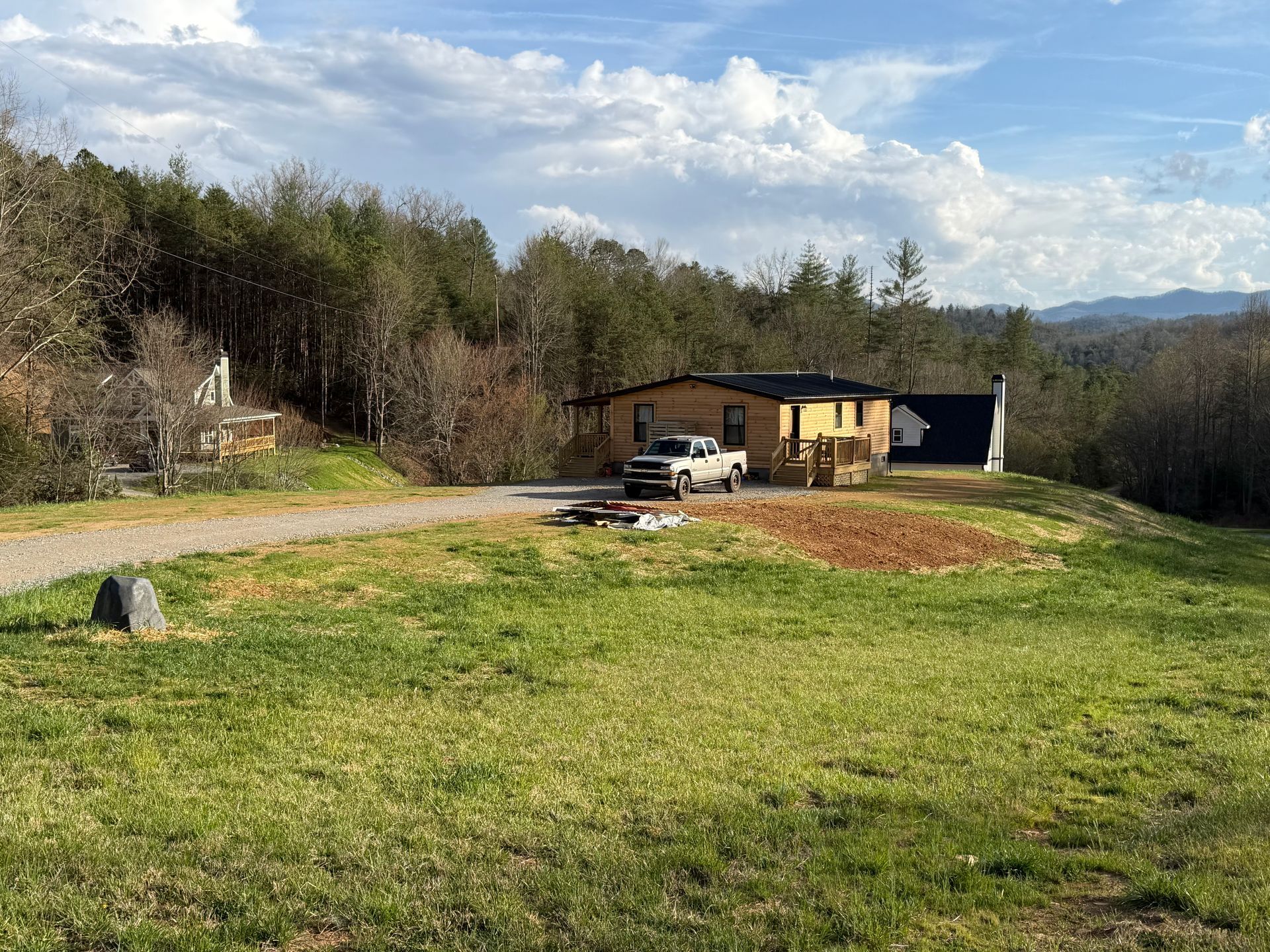 Rural cabin with a parked pickup on a gravel drive, surrounded by grass and trees under a blue sky.