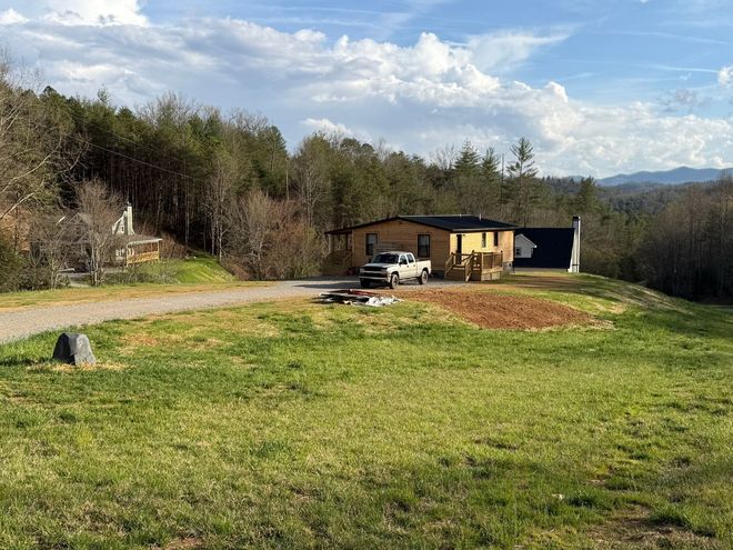 Rural cabin with parked pickup truck beside a gravel drive and open grassy field under a blue sky