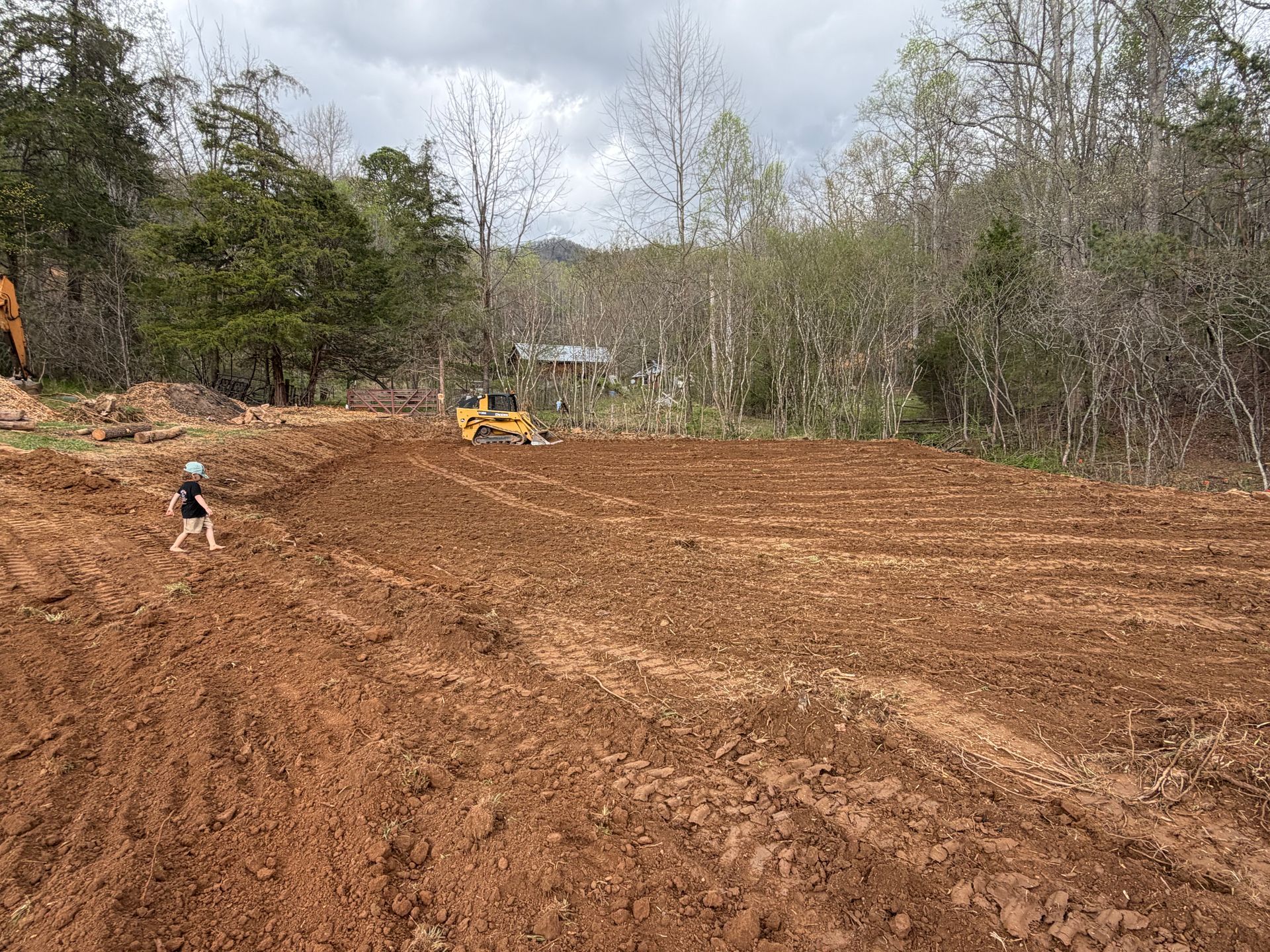 Cleared dirt lot with a small excavator, muddy tracks, and trees in the background