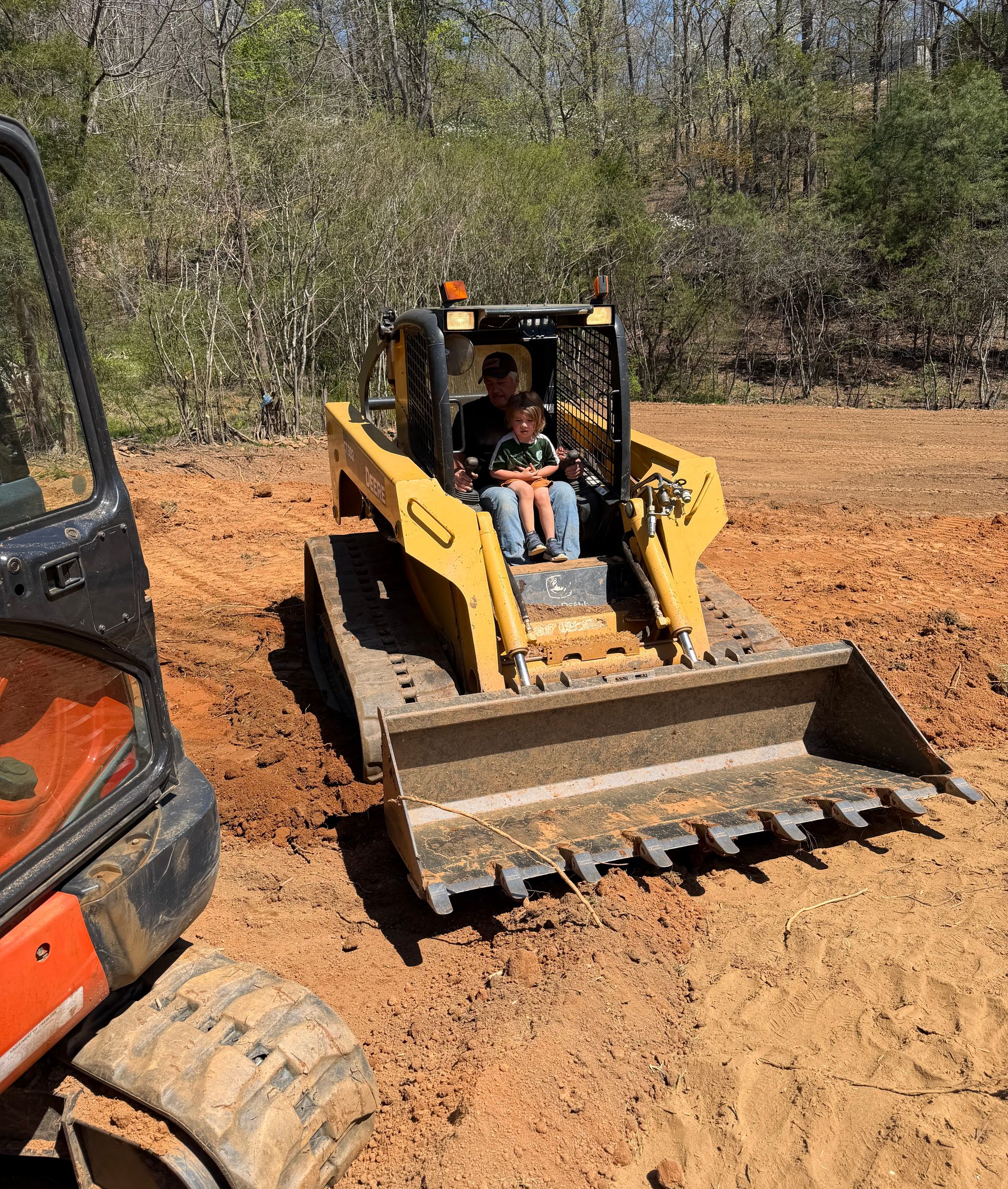 Yellow skid steer with front loader on a dirt lot, carrying two people in the cab.