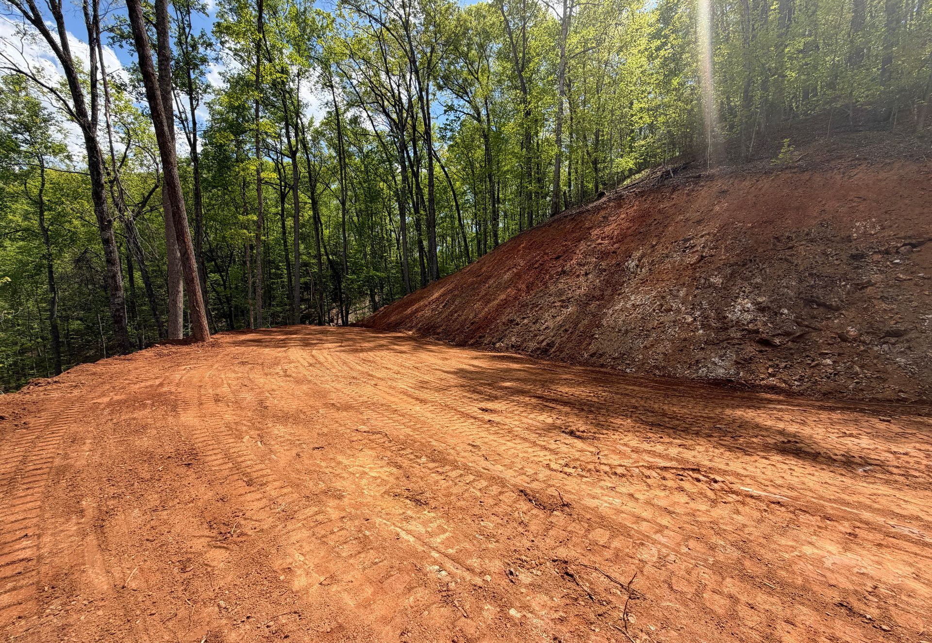 Freshly graded red dirt road beside a wooded hillside under bright sunlight