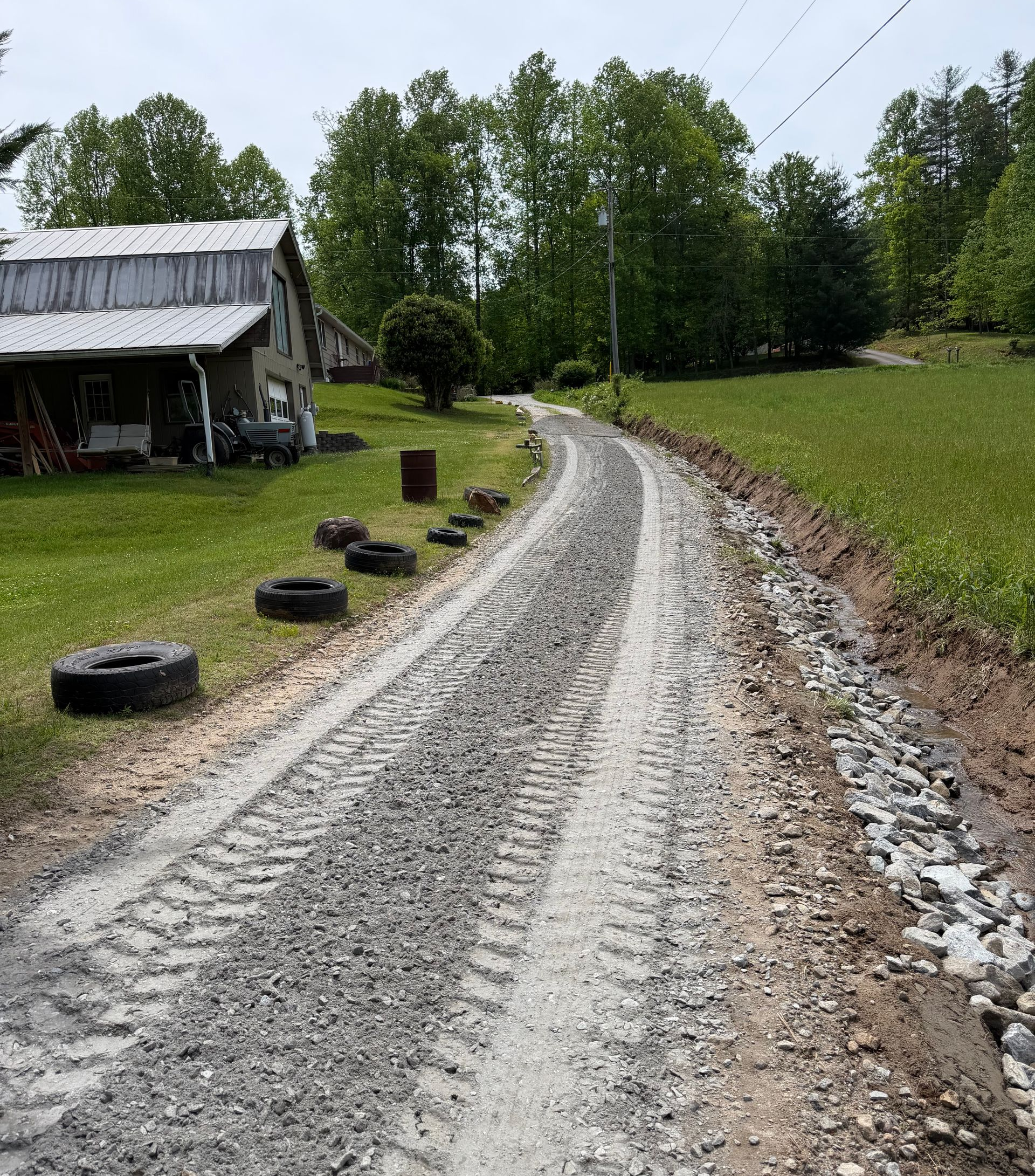 Gravel road curving past a rustic house and tire-lined yard in a green rural landscape