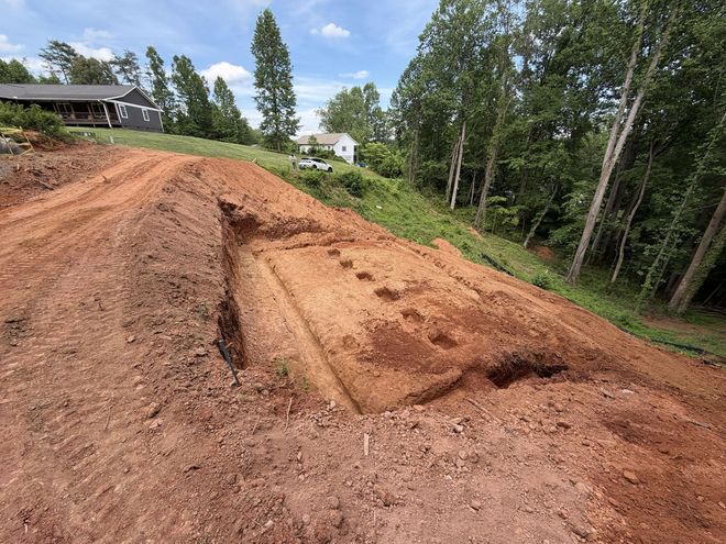Dirt slope with a carved trench on a wooded hillside under a partly cloudy sky