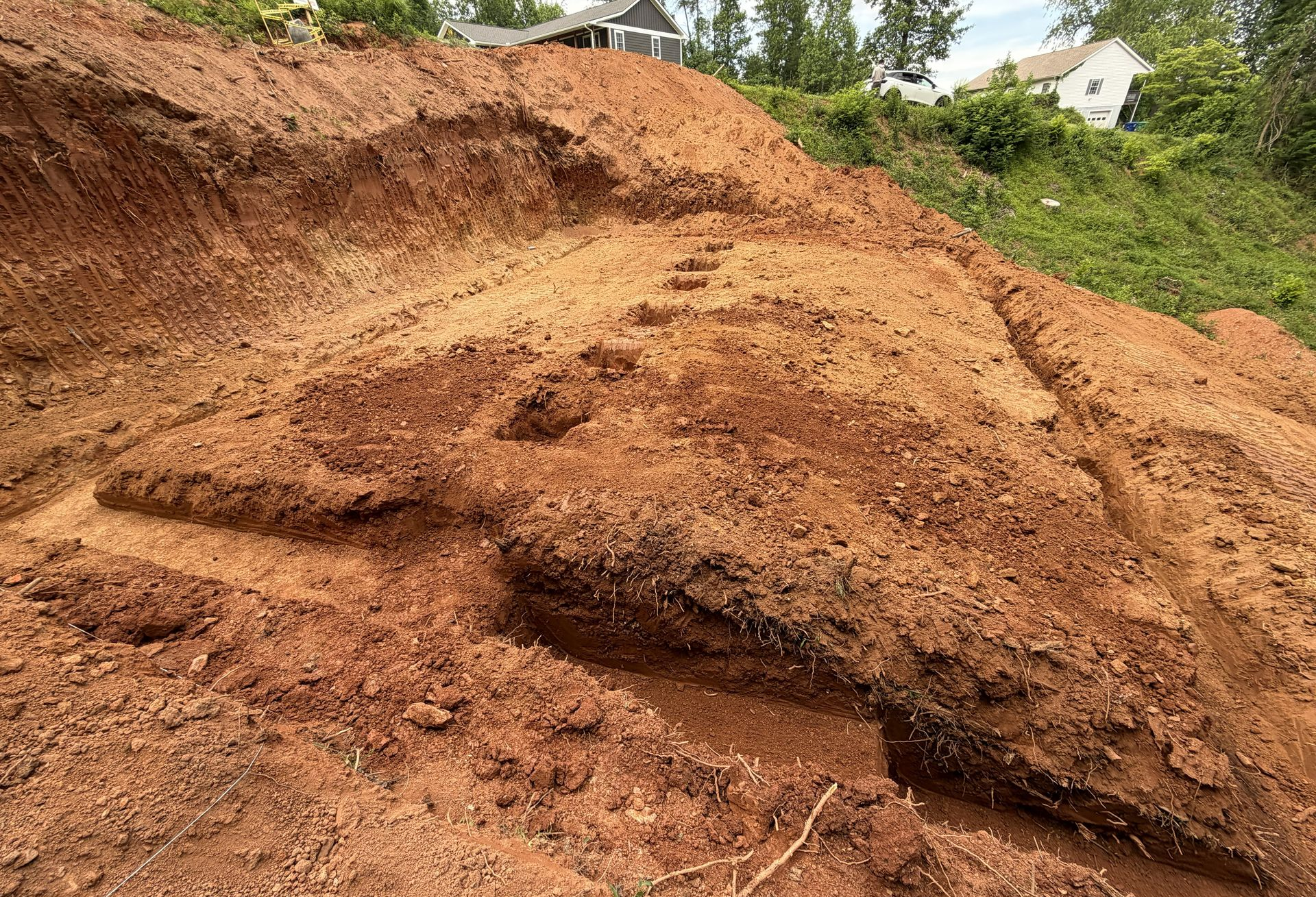 Large reddish-brown dirt landslide on a hillside with vegetation and a few houses in the background