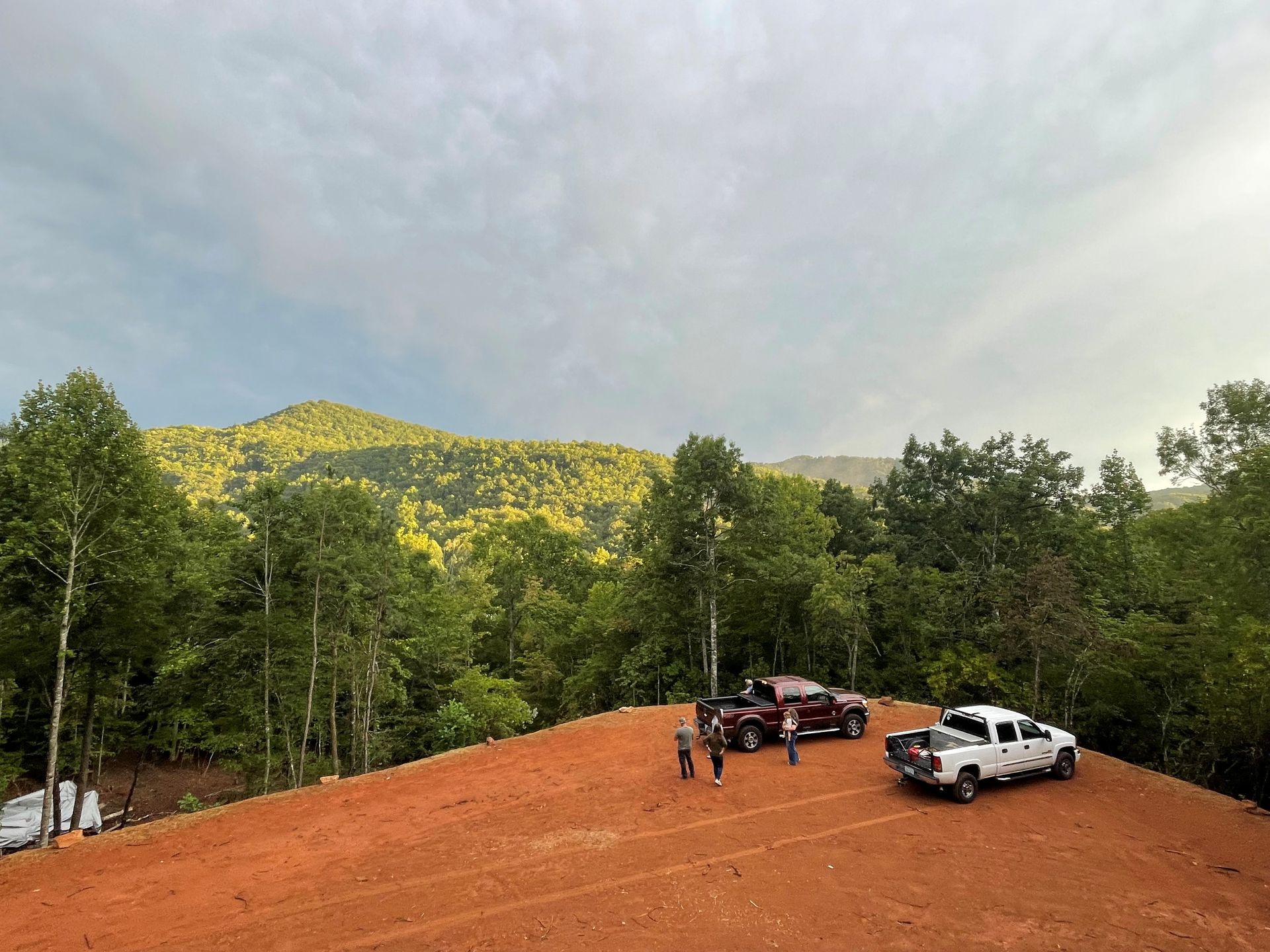 Parked trucks on a red dirt clearing beside a forested hillside under a cloudy sky