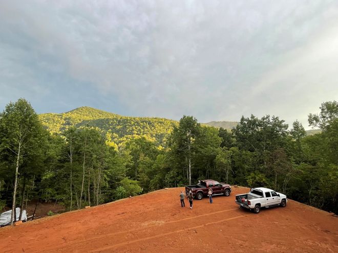 Parked trucks on a red dirt clearing beside a forested hillside under a cloudy sky