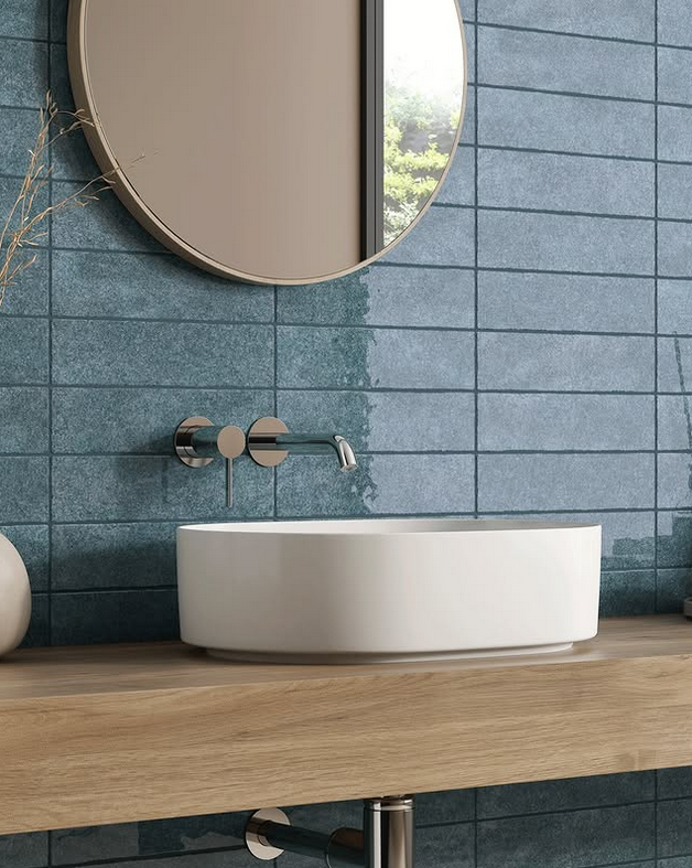 Bathroom with blue tiled wall, white sink, wooden counter, and round mirror.