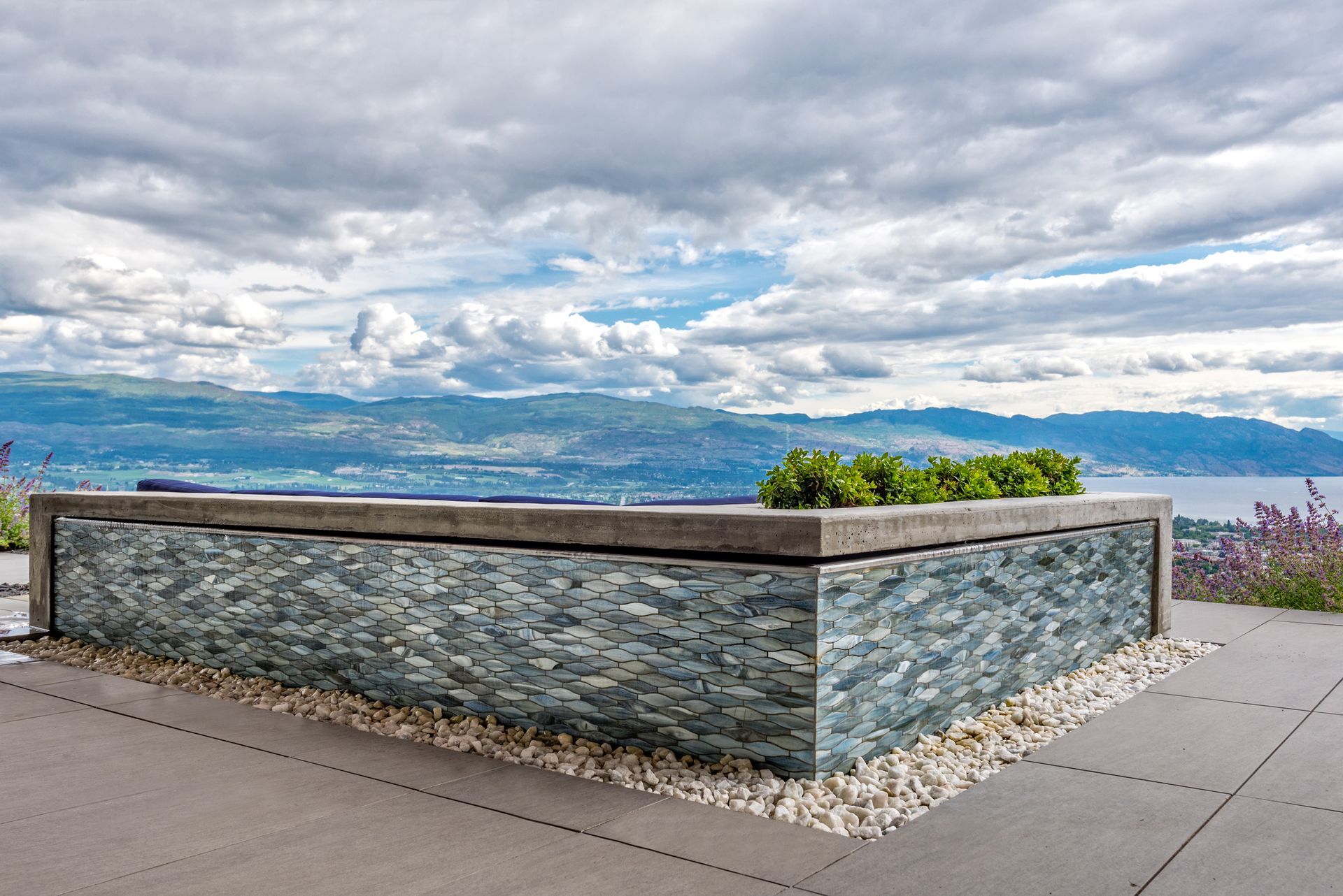 Hot tub overlooking a scenic mountain and lake view, with stone exterior and cloudy sky.