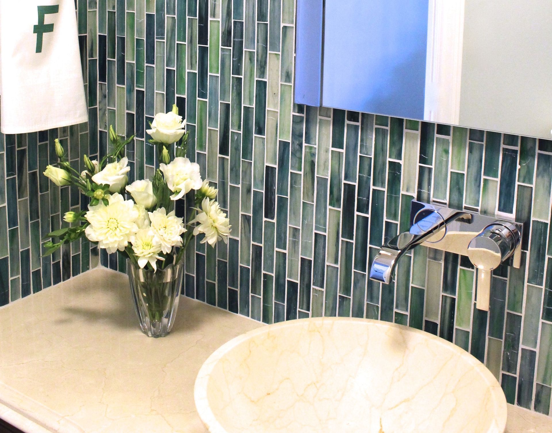 Bathroom with green mosaic tile backsplash, cream countertop, light wood sink, and white flowers.