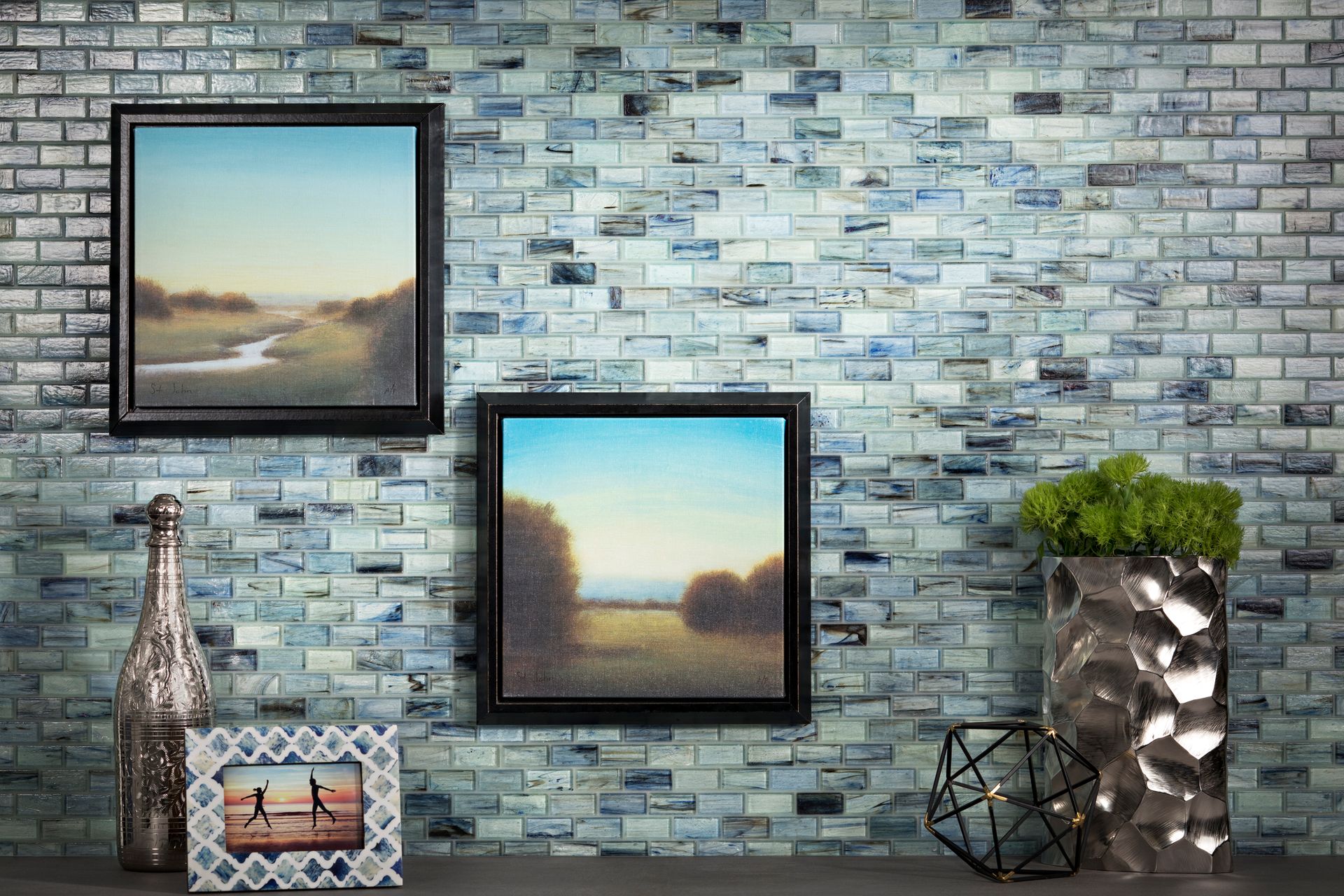 Two framed landscape paintings and decorative objects on a shelf against a blue brick wall.