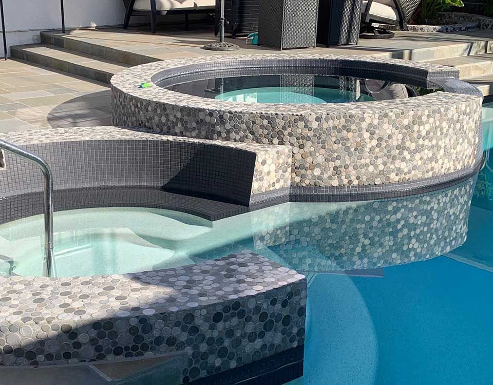 Pool with a built-in hot tub, surrounded by mosaic tile, steps, and patio furniture.