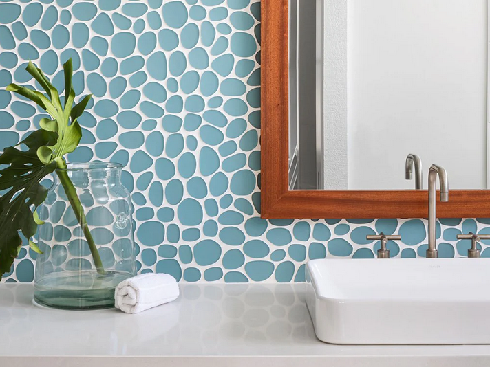 Bathroom with blue pebble tile backsplash, white sink, wooden mirror frame, and plant in a glass vase.