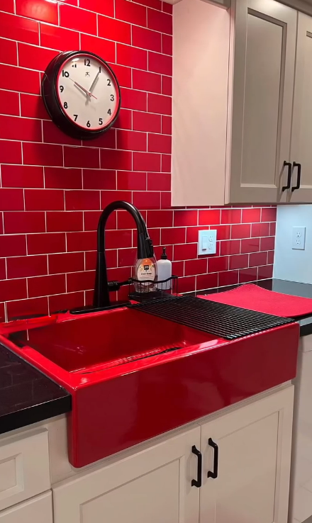 Red kitchen sink and backsplash with a black faucet. White cabinets and clock.