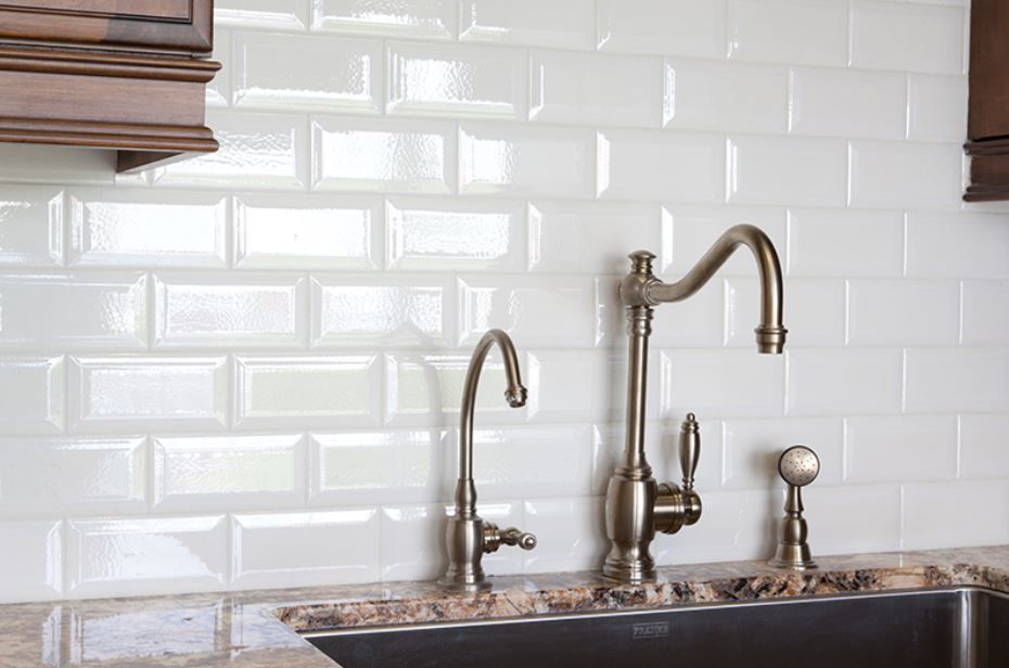 White subway tile backsplash with bronze faucets over a stainless steel sink.