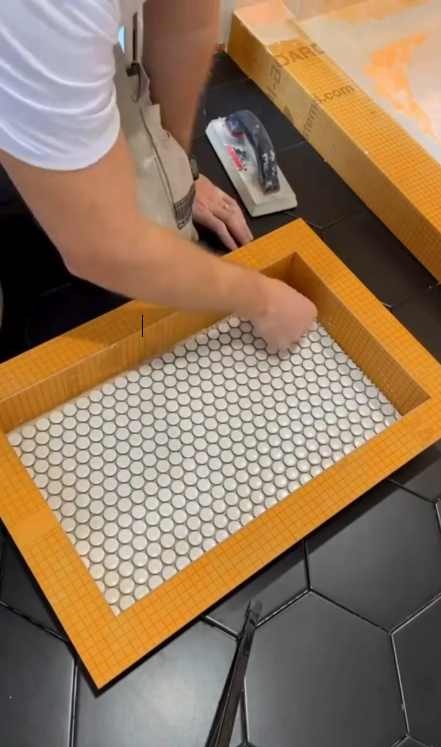 Man installing small, white mosaic tile in a rectangular shower niche frame, with a black hexagon tile floor.