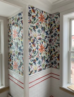 Colorful floral wallpaper adorns a corner, above white tile with red stripes. Two windows flank the walls.
