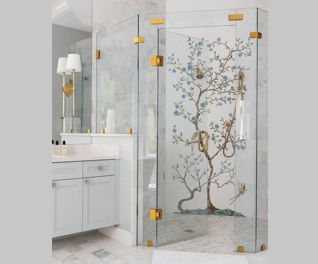Glass shower with painted tree design and gold hardware in a white bathroom.