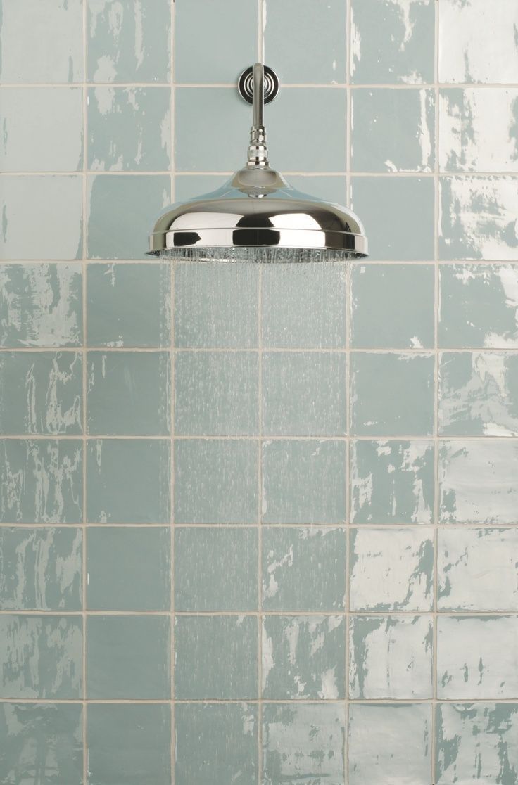 Shiny chrome shower head with water spraying, mounted on blue-green tiled wall.