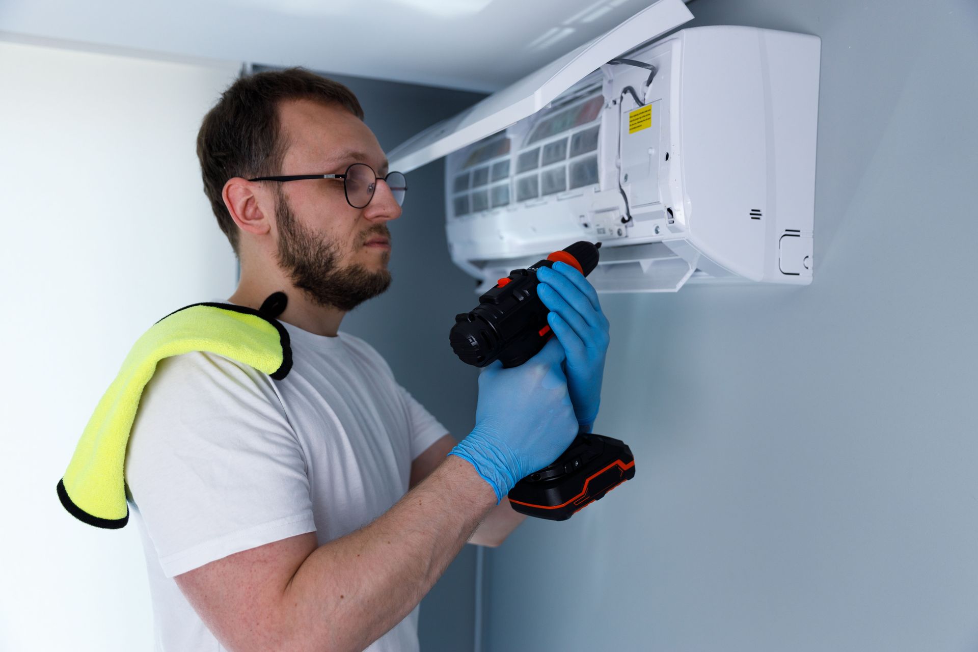 Man in blue gloves using a drill to install or repair a wall-mounted air conditioner.