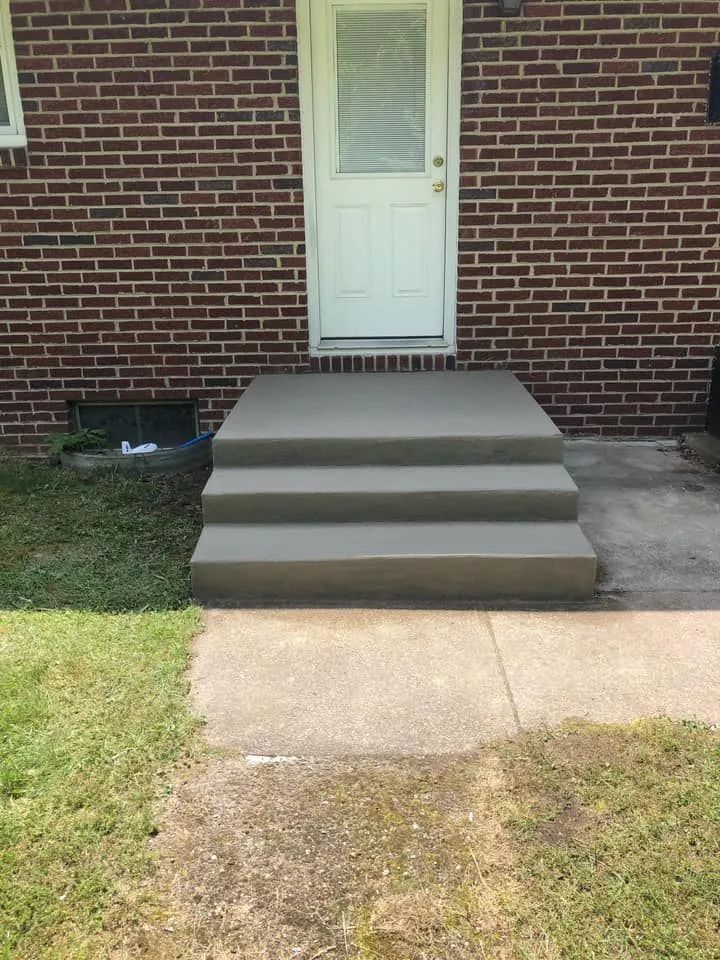 A set of three new, smooth grey concrete steps leading up to a white door on a red brick house exterior.