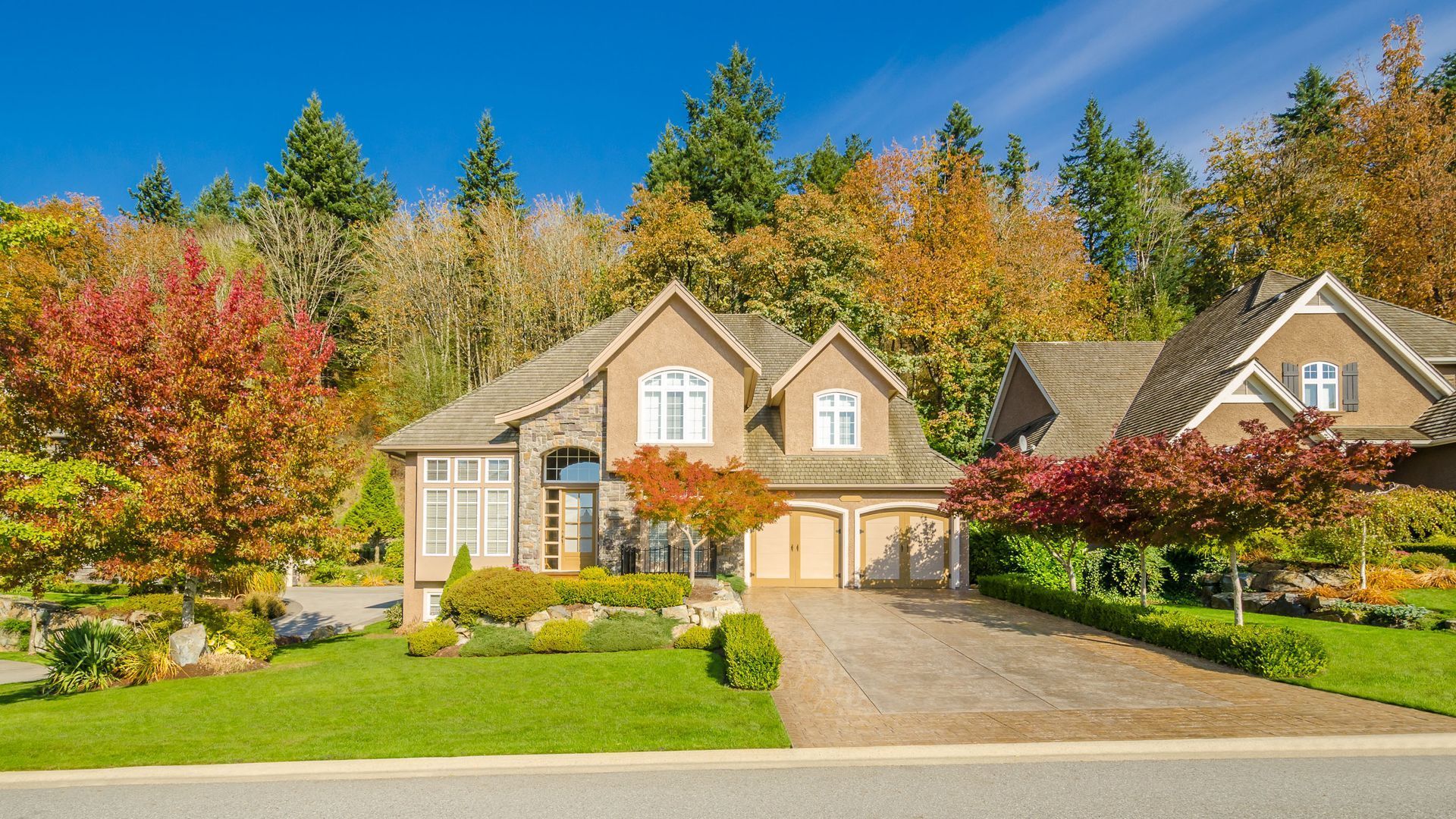 A tan house with a paved driveway and green lawn sits against a backdrop of colorful autumn trees under a bright blue sky.