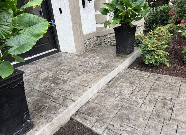 A paved porch and walkway with textured, grey stone-patterned concrete, framed by black planters and landscaping.