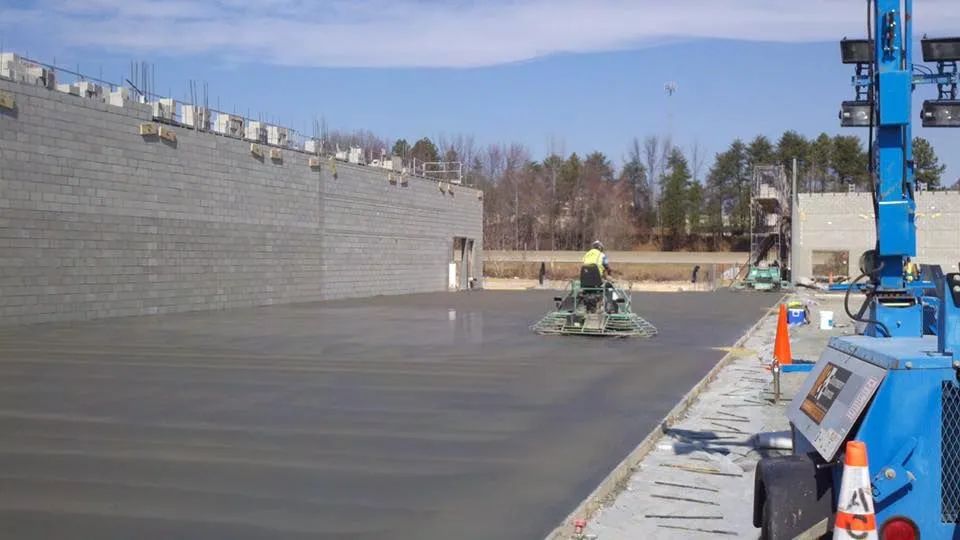 A person operating a ride-on power trowel to smooth wet concrete at an outdoor construction site with a long brick wall.