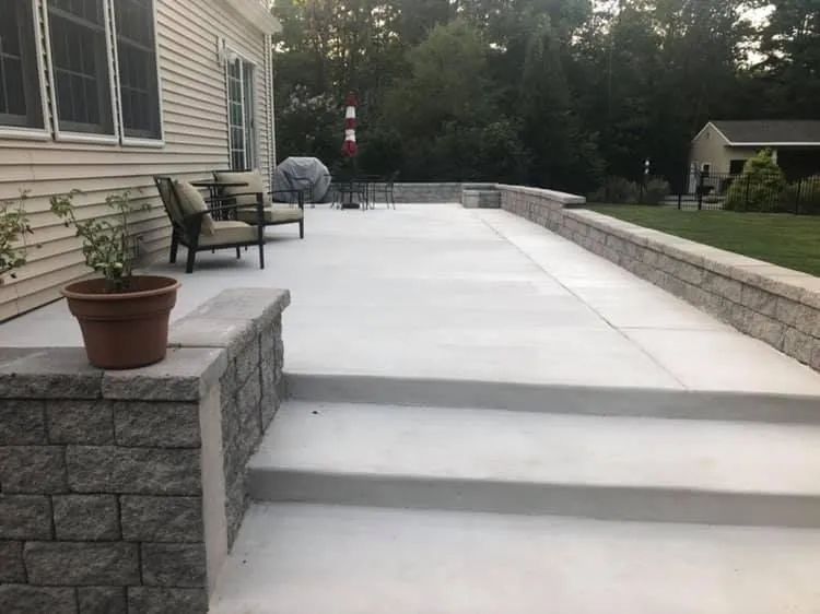 A patio with light grey concrete steps, a stone retaining wall, two chairs, and a potted plant against a house exterior.