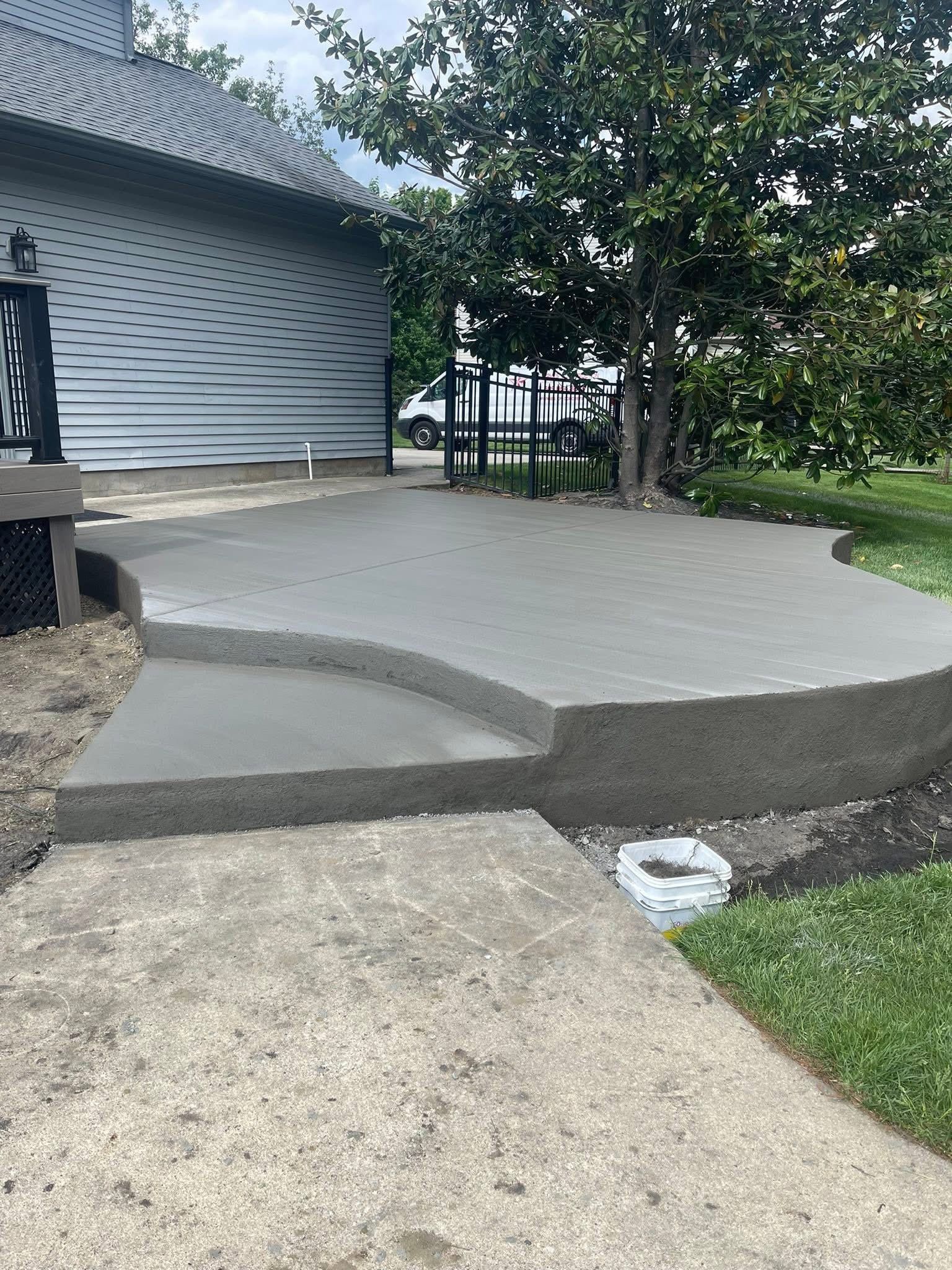 A newly poured concrete patio with a single step, located outside a building near a tree.