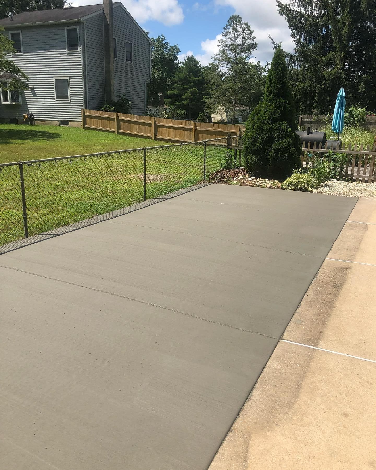 A freshly poured concrete patio next to a chain-link fence, a wooden fence, and a house in a residential backyard.