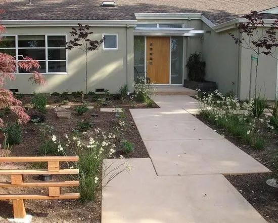 A modern, single-story home with light grey siding, a wood-paneled door, and a sidewalk leading through a garden.