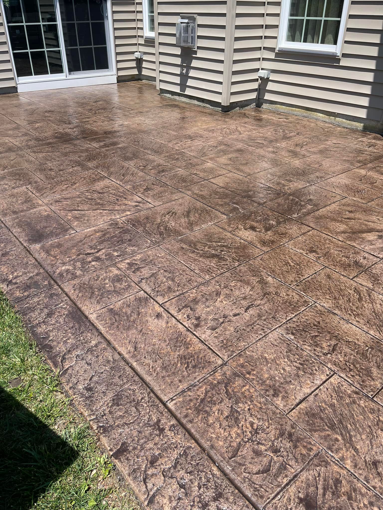 A patio with stamped concrete that resembles brown stone tiles, positioned next to a house with beige siding.