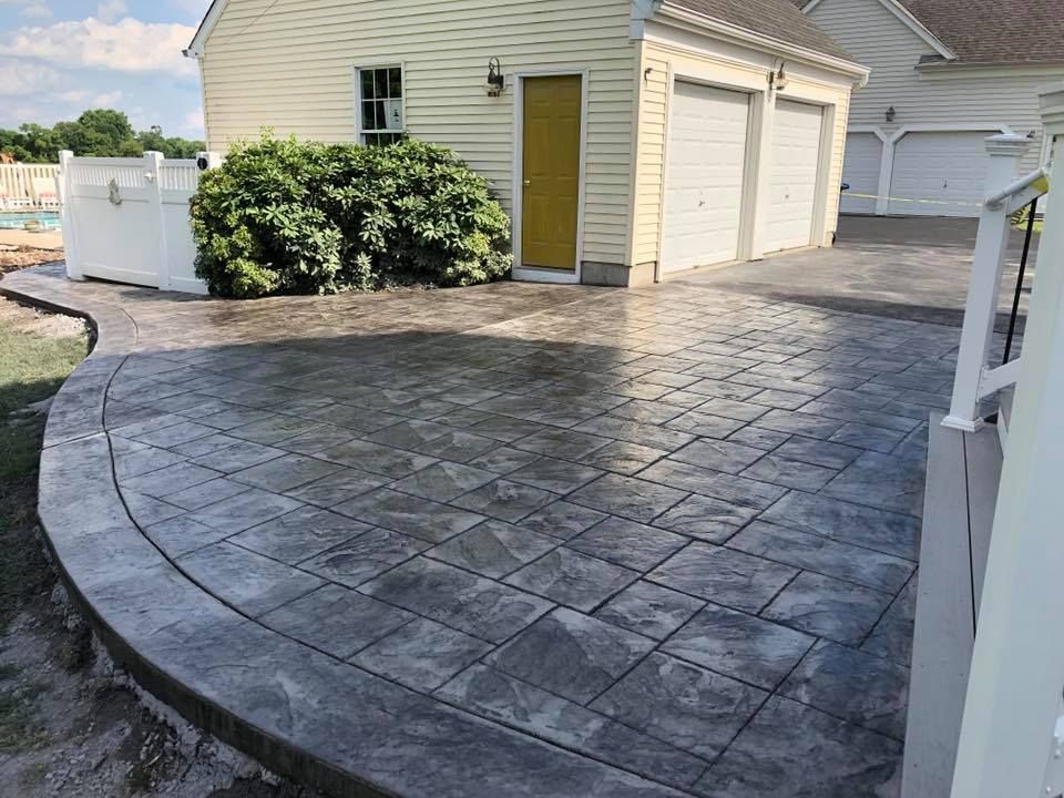 A dark gray stamped concrete patio with a rectangular tile pattern in front of a light-sided house with a garage.