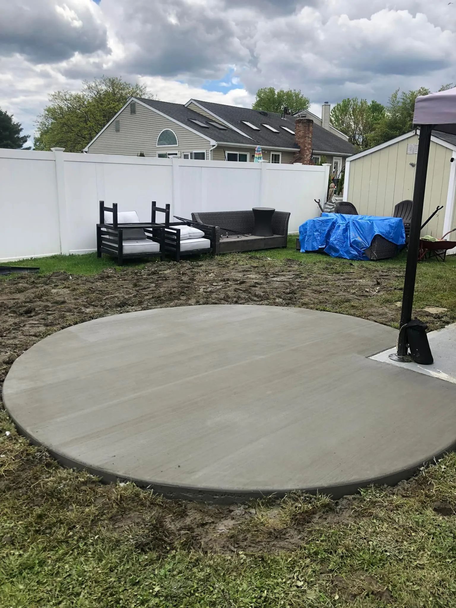 A new circular concrete patio sits in a backyard next to white fencing, patio furniture, and a blue tarp-covered object.