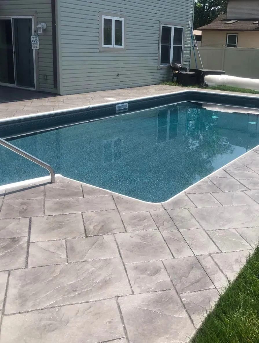 An rectangular swimming pool with deep blue water, surrounded by grey stone patio tiles next to a green-sided house.