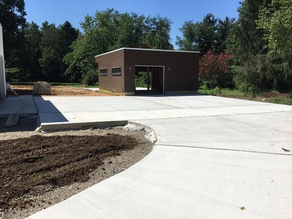 A newly poured concrete driveway leads to a small, dark-brown detached garage on a sunny day with trees in the background.
