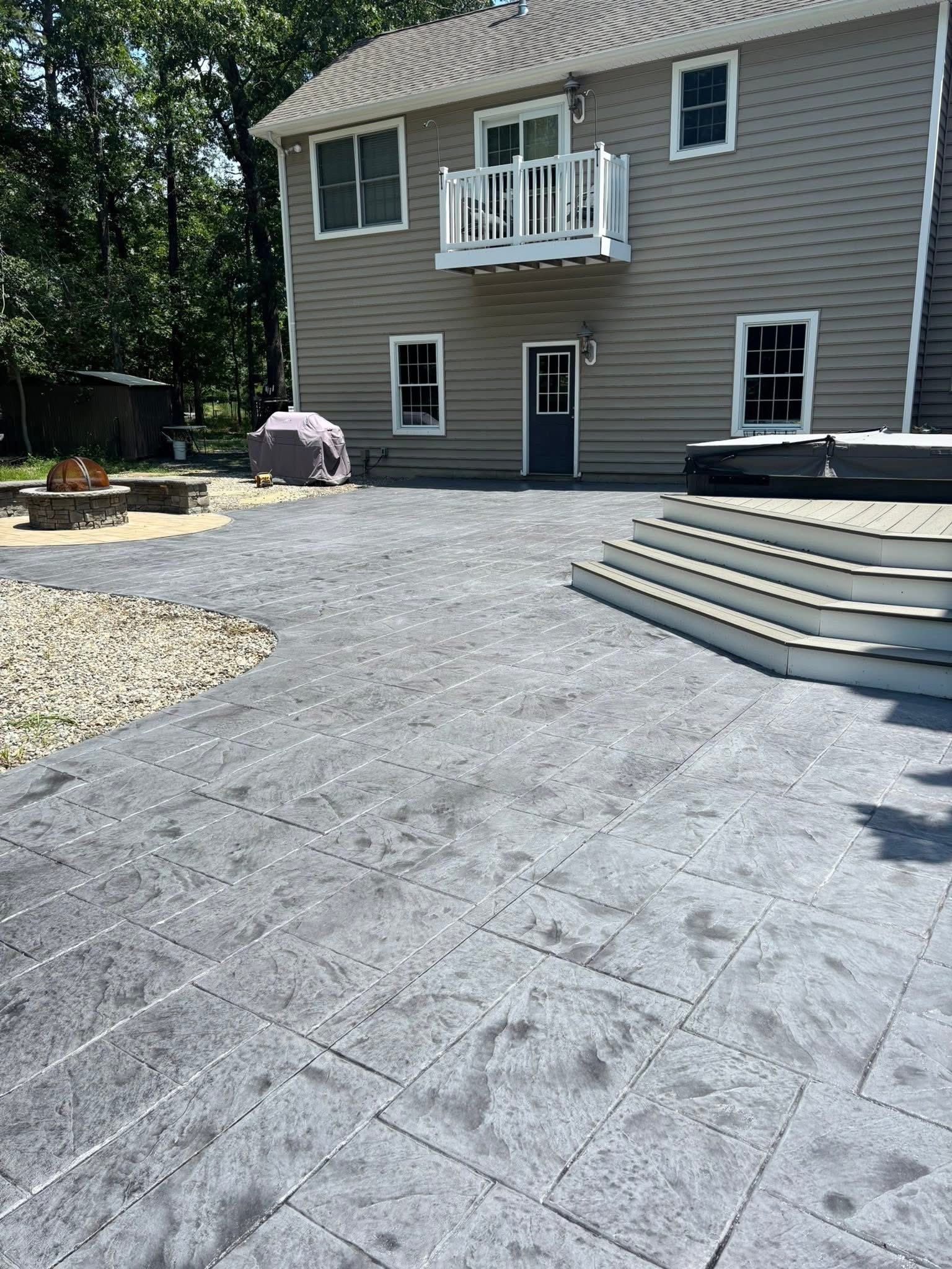 A beige, two-story house with a grey stone-patterned patio, stairs leading to a hot tub, and a small fire pit area.