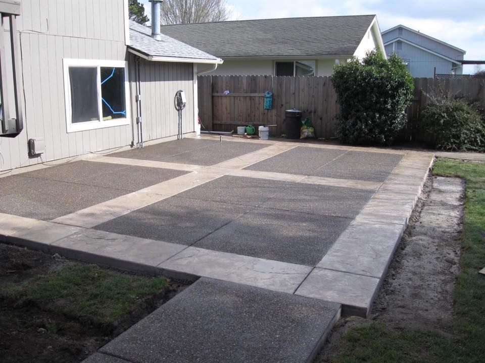 A newly poured concrete patio with a rectangular grid design sits in a backyard next to the side of a grey house.