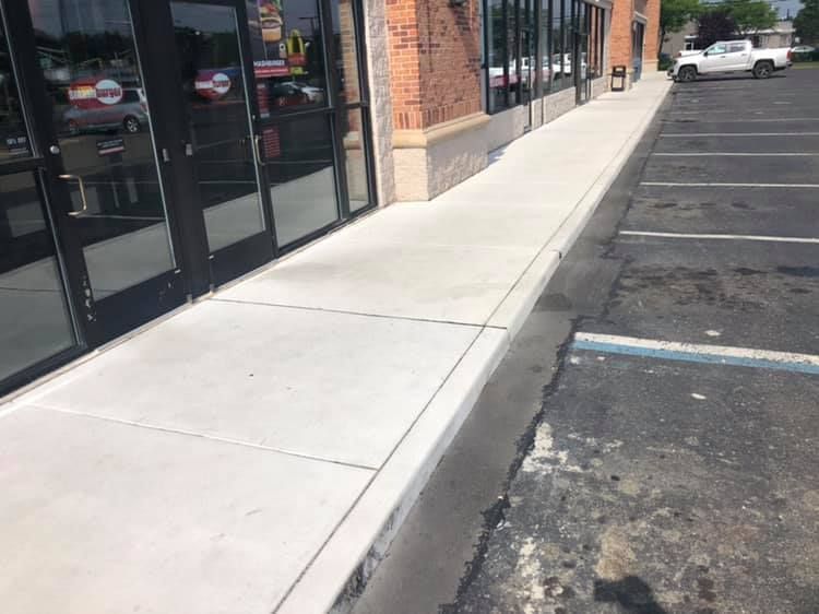 A newly poured concrete sidewalk runs along a brick storefront, adjacent to an asphalt parking lot with painted lines.
