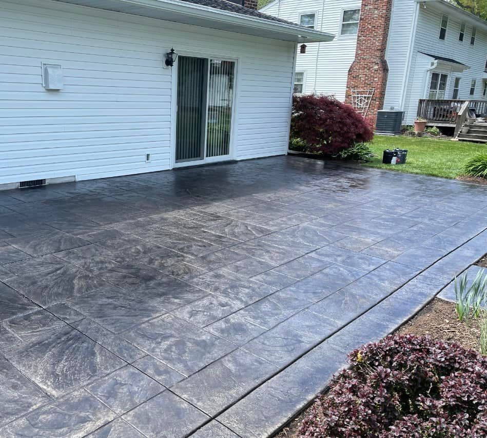A dark gray stamped concrete patio outside a white house with a sliding glass door and a small bush nearby.