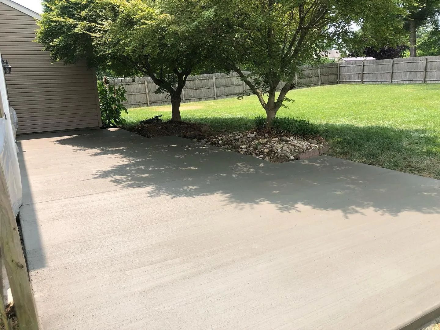 A newly poured gray concrete patio sits beside a house wall, shaded by two trees with a lawn and fence in the background.