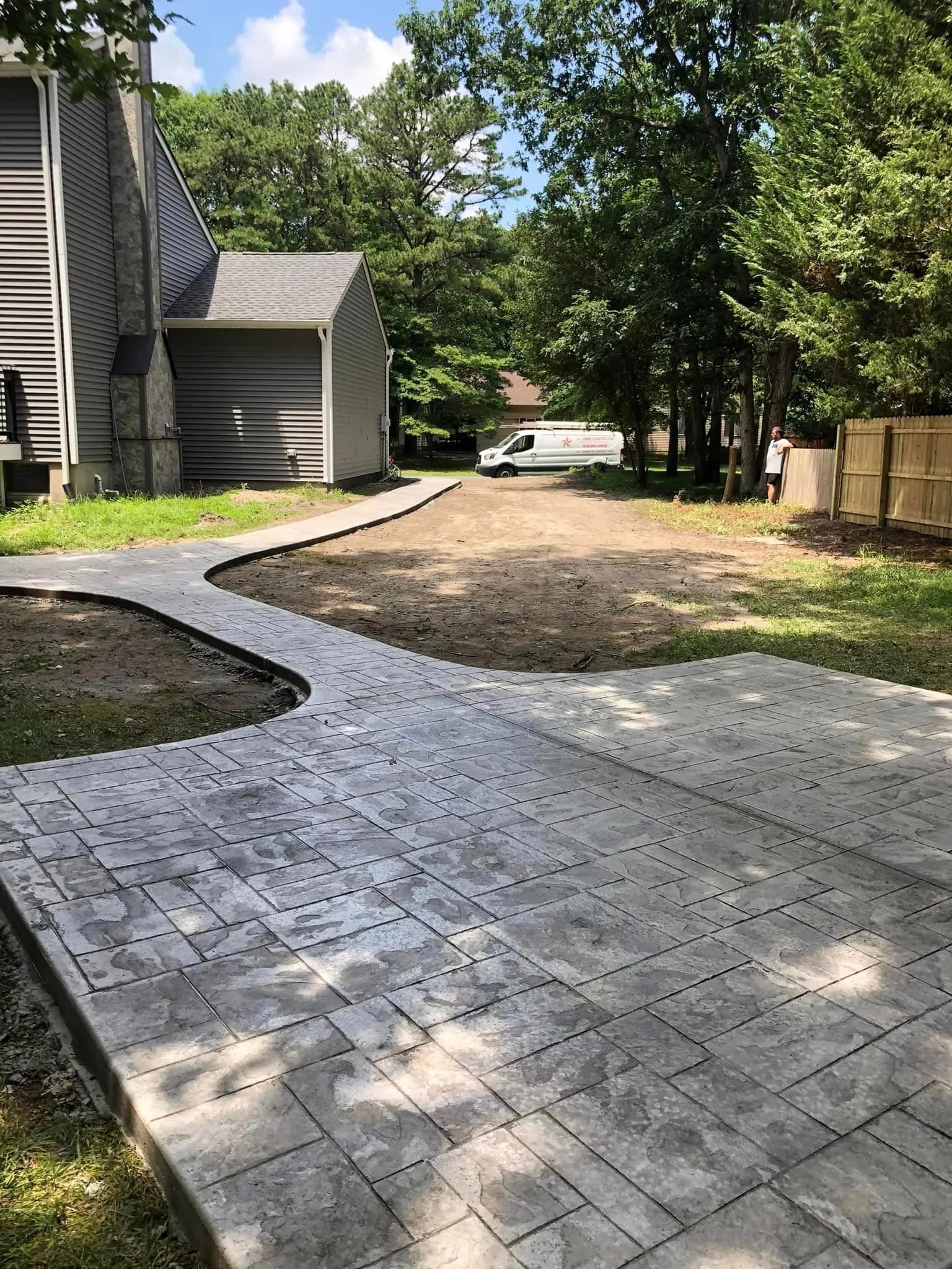 A wide-angle view of a newly installed gray stone paver patio and walkway extending from a house toward a wooded backyard.