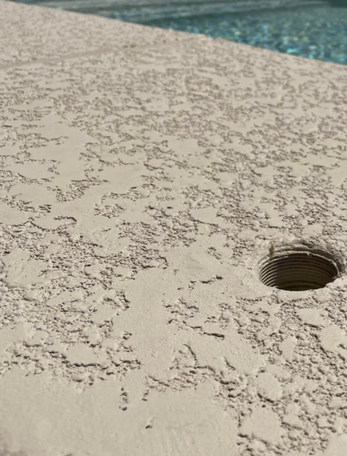 A close-up of a beige, textured pool deck surface featuring a single circular hole with visible threads.