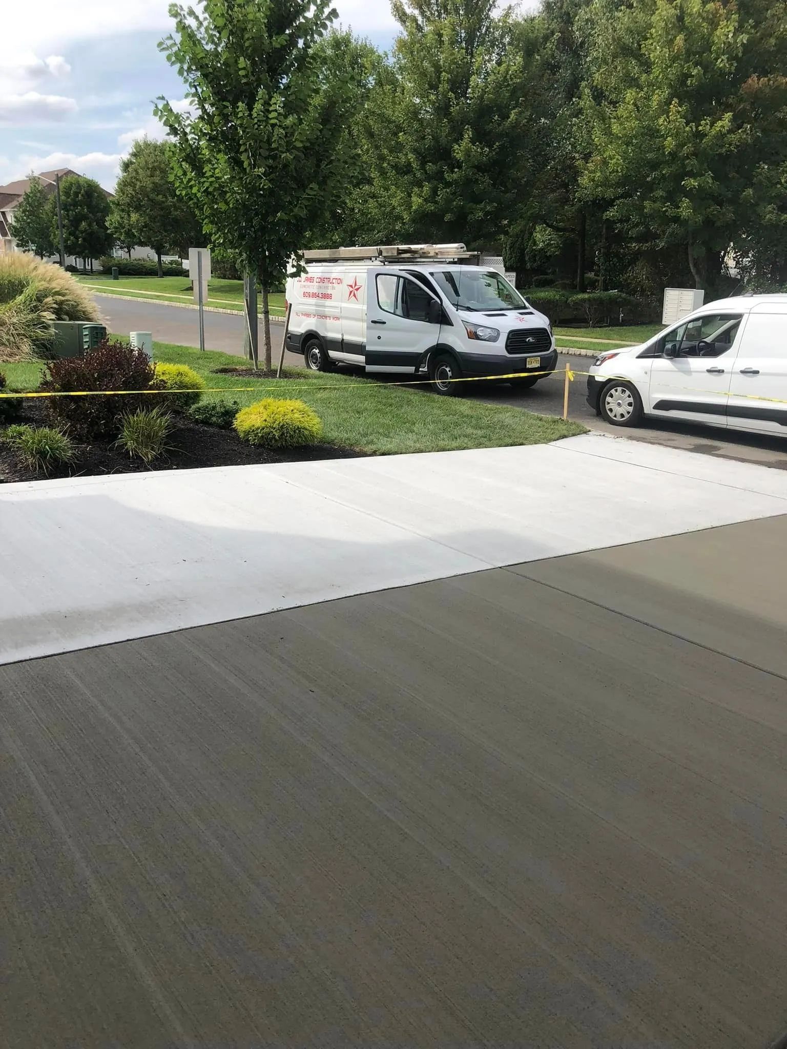 Two white service vans parked on the grass beside a freshly poured concrete driveway on a sunny day.