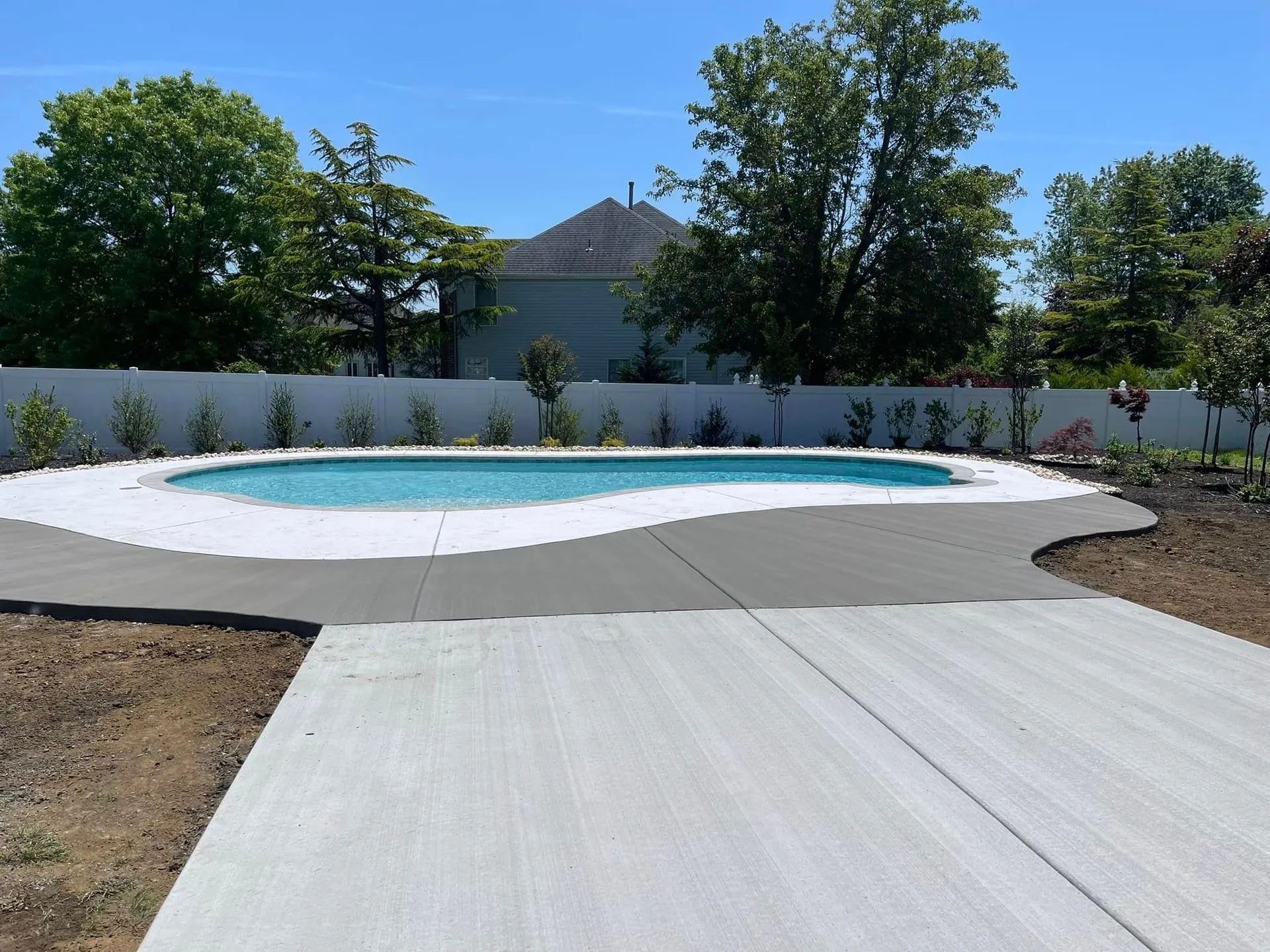 A backyard swimming pool with light blue water surrounded by a new, grey concrete patio against a white vinyl fence.