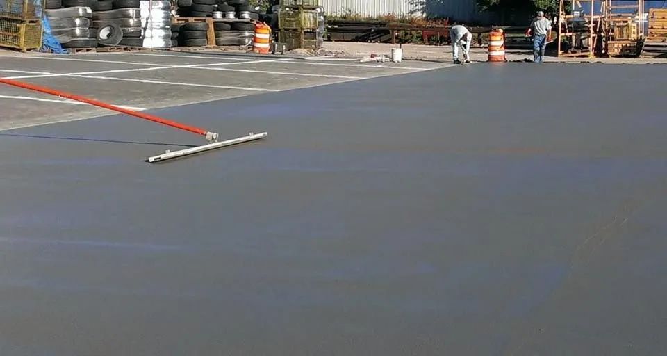 A worker uses a concrete rake to smooth a freshly poured, wet gray concrete slab in an outdoor construction area.